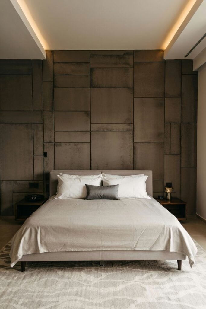 A modern bedroom wall is composed of large, irregular rectangular concrete panels. Linear lighting illuminates the top edge of the wall. A simple bed with neutral bedding is placed in front of the dark-toned wall, with minimalist nightstands and lamps on either side. A light-colored rug is on the floor.