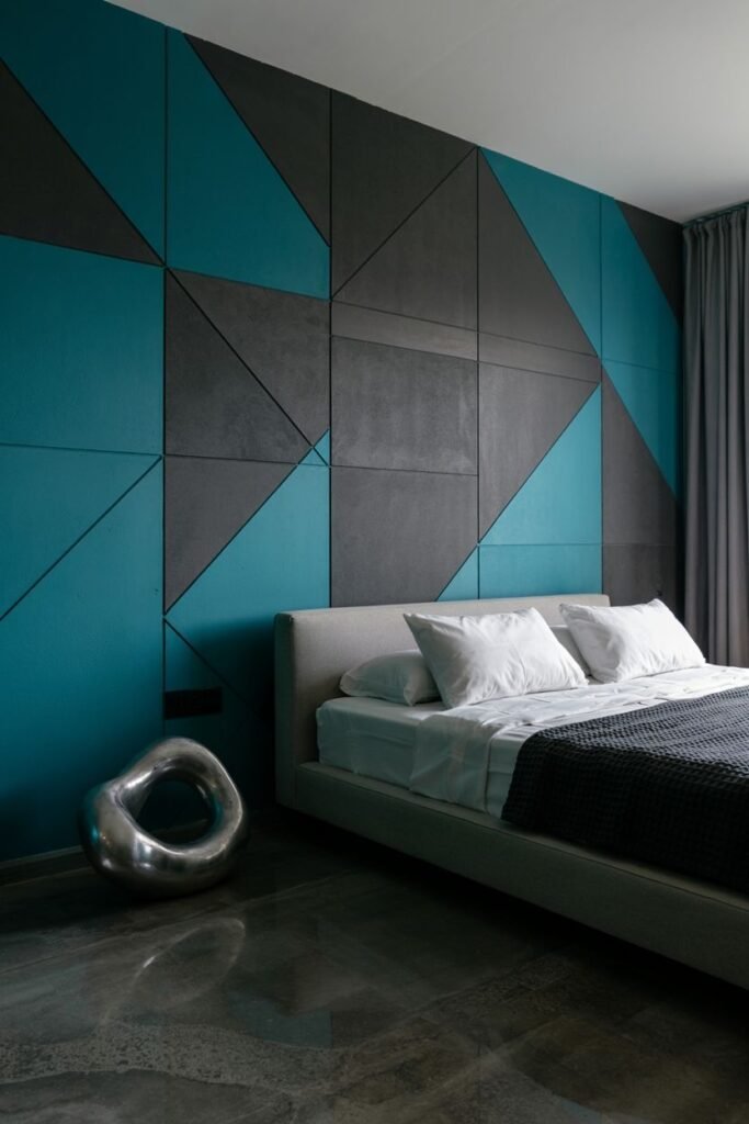 An accent wall features large geometric shapes in contrasting colors of black, grey, and teal. A simple bed with white bedding is placed against the wall. A modern silver sculpture is on the floor. A polished concrete floor is visible.