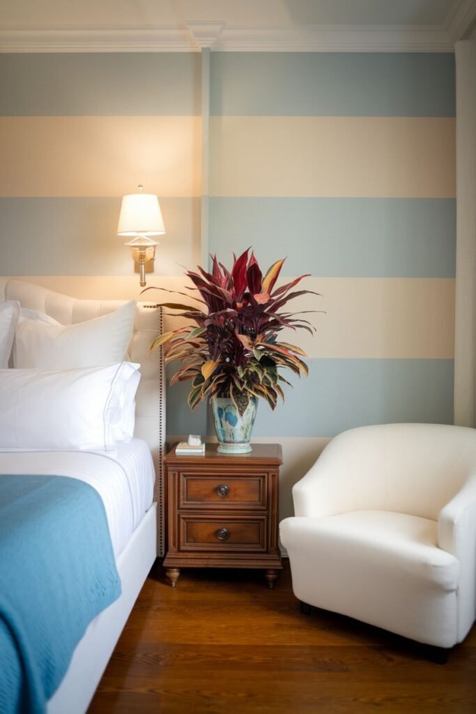A plant with red and yellow variegated leaves is displayed in a large, patterned blue and white urn on a wooden nightstand next to an armchair.