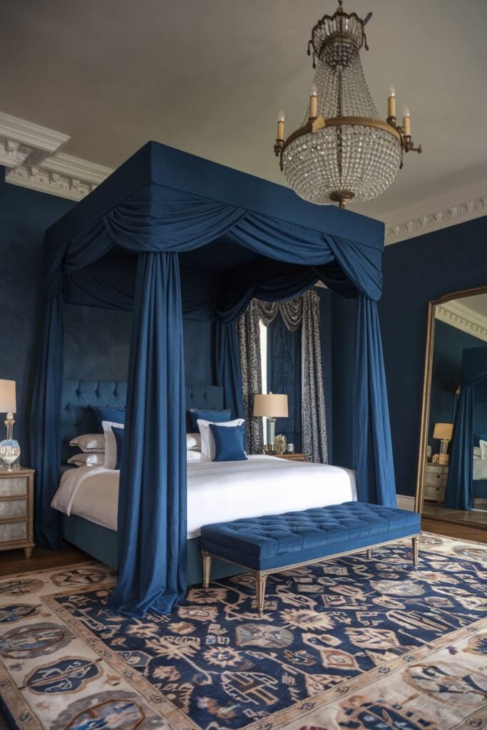 A dramatic royal bedroom featuring deep blue walls and a coordinating blue four-poster bed with draped fabric, a tufted bench, a large mirror, a crystal chandelier, and a comfortable patterned rug.