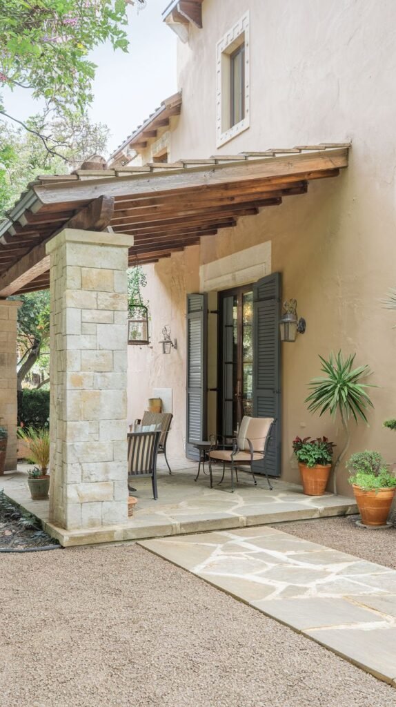A covered patio with a stone slab floor and a stone column supporting the roof. The house wall is stucco, and there are dark-colored shutters on the windows. The patio is furnished with outdoor seating. A gravel path leads to the patio.