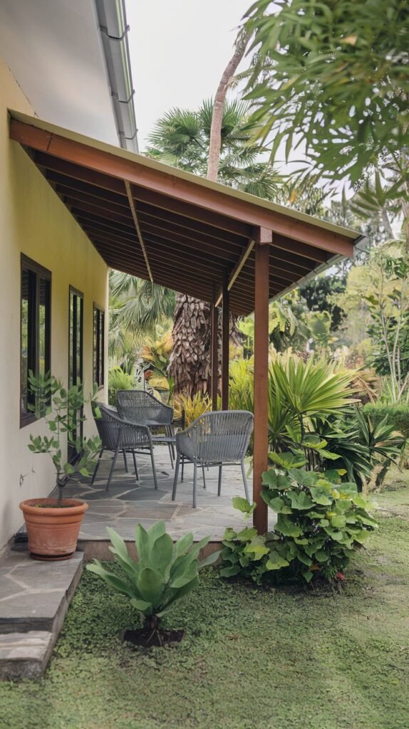 A covered patio with a stone tile floor surrounded by lush tropical plants. The patio is furnished with grey outdoor chairs and a table. The roof is supported by wooden posts.