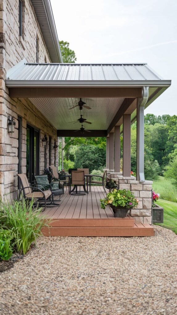 A covered wooden deck porch with a metal roof, attached to a stone house. The deck is furnished with outdoor chairs and a dining set, and ceiling fans are installed in the roof. Gravel is in the foreground.