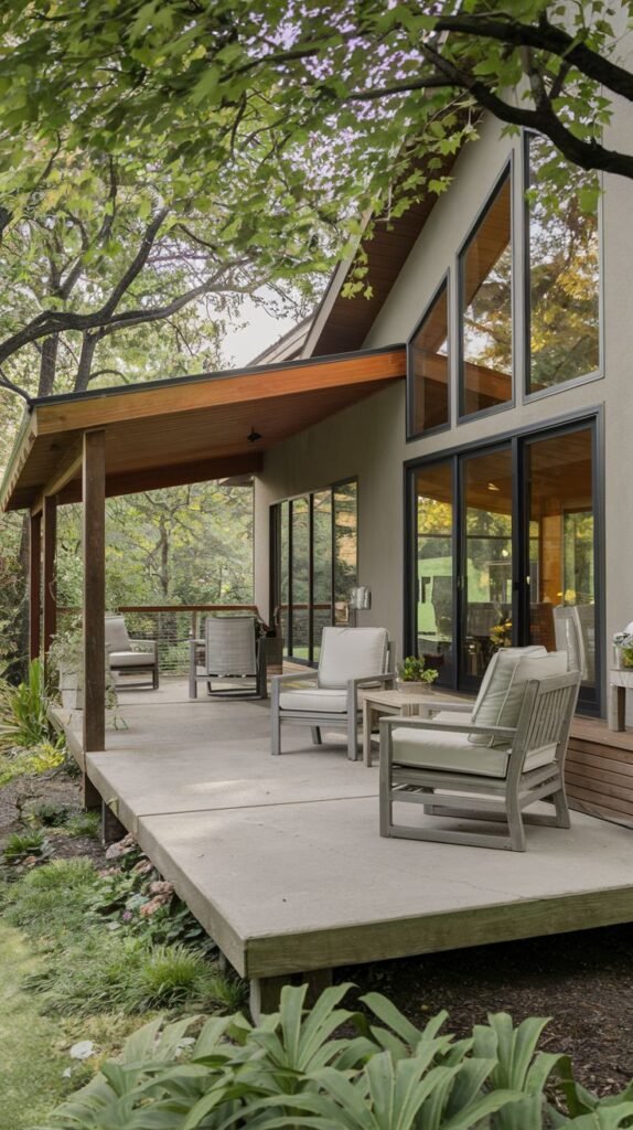 A covered patio with a concrete deck. The patio is furnished with modern outdoor chairs and a side table. The roof is supported by a wooden structure, and the house wall has large windows and a sloped roofline.