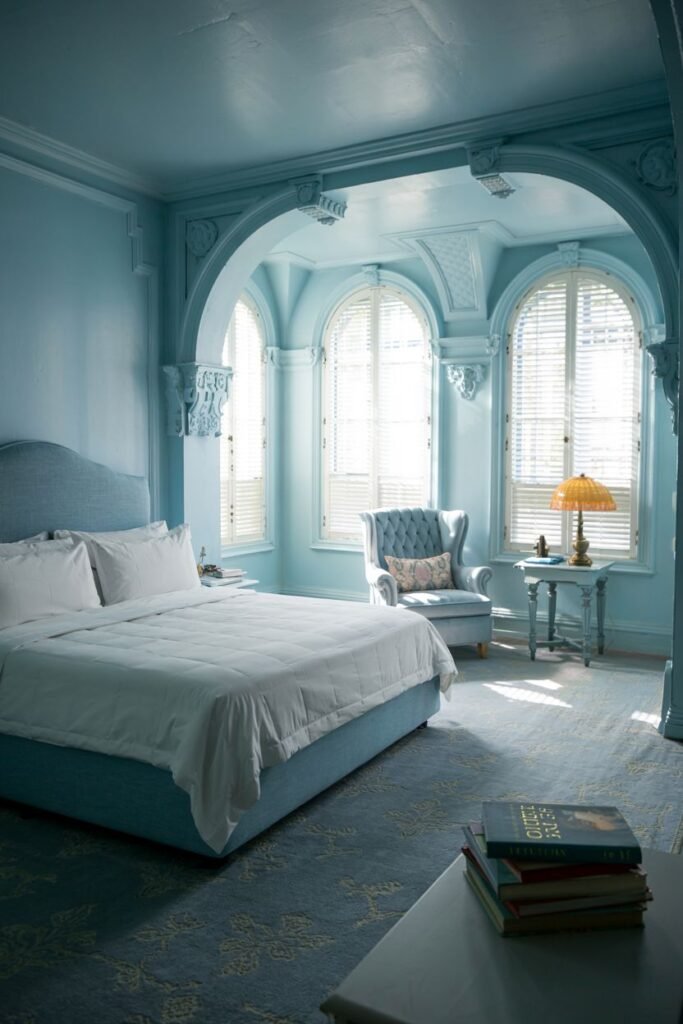 A unique and cozy bedroom with light blue walls and architectural arched window frames with blinds, featuring a bed and armchair.