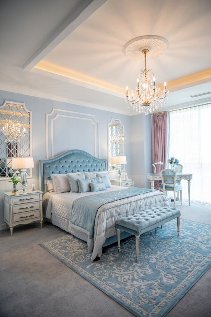 A bright and cozy bedroom featuring light blue walls and a tufted blue headboard, with a crystal chandelier and a patterned rug.