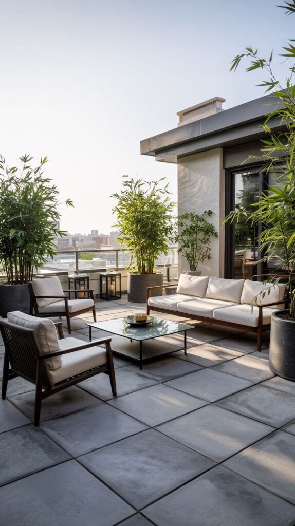 A modern and cozy rooftop terrace with a seating area featuring a sofa and armchairs around a glass coffee table, surrounded by large potted bamboo plants against a city skyline background.