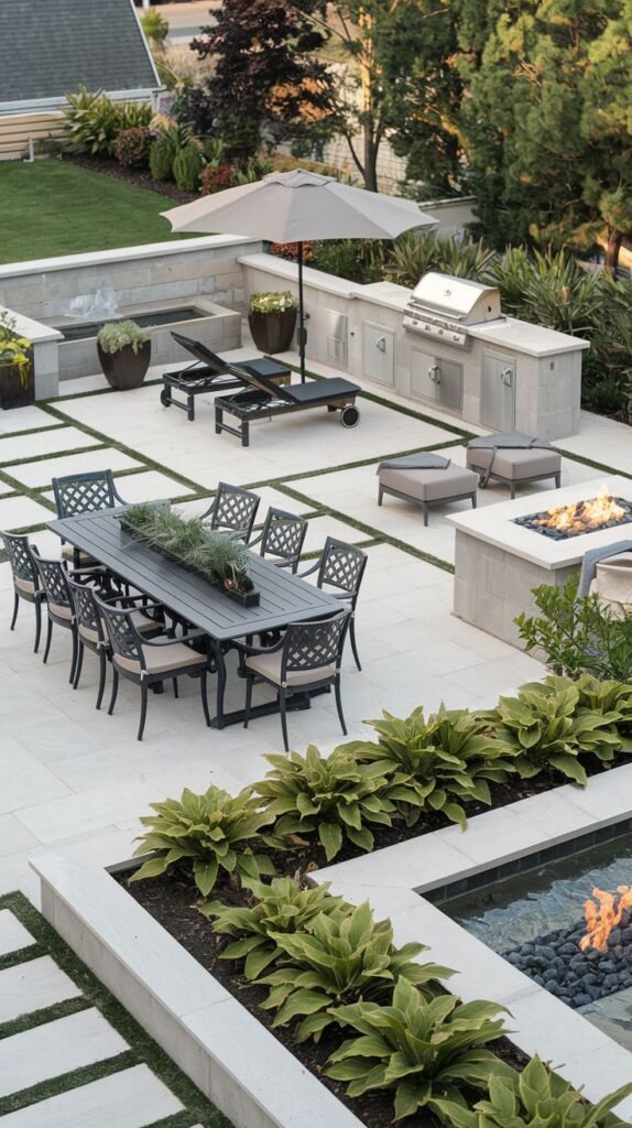 A large, modern backyard patio designed for entertaining, featuring a dining area, lounge chairs, a fire pit area, and a built-in grill/kitchenette, all set on large light-colored pavers with integrated planting beds and a water feature, offering a cozy resort-like feel.