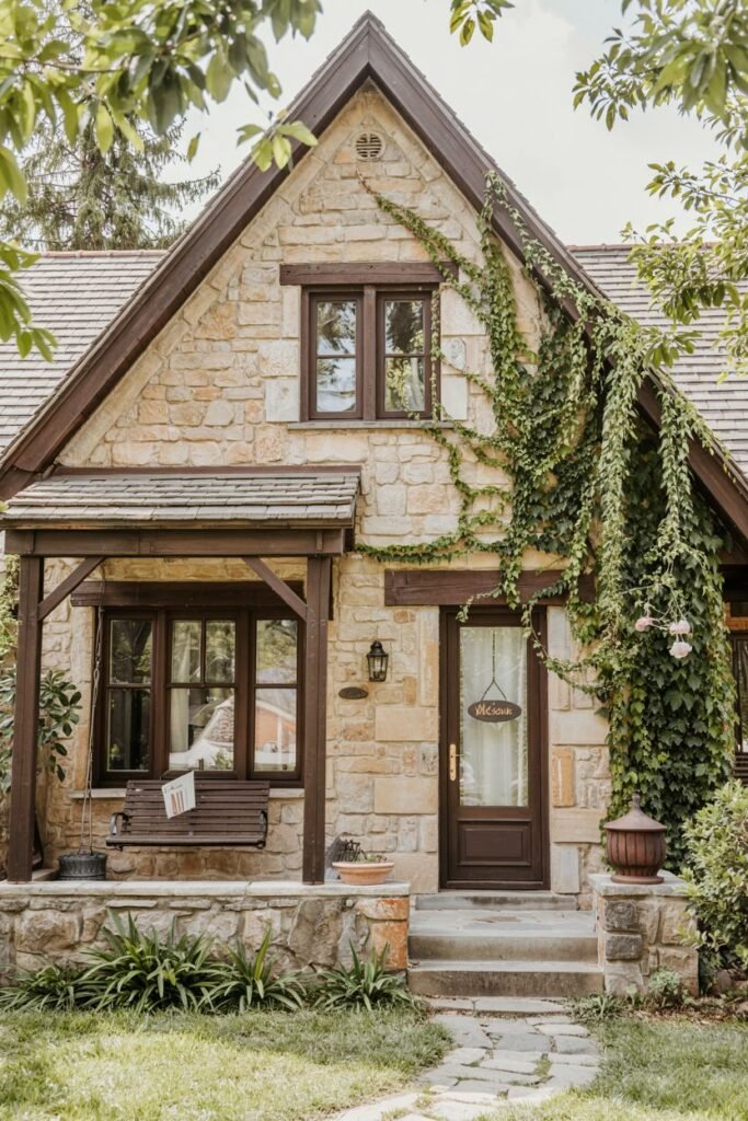 A small, rustic stone cottage covered in climbing ivy, with a welcoming porch swing and a sign that says "Welcome". The warm lighting from the interior suggests a cozy atmosphere.