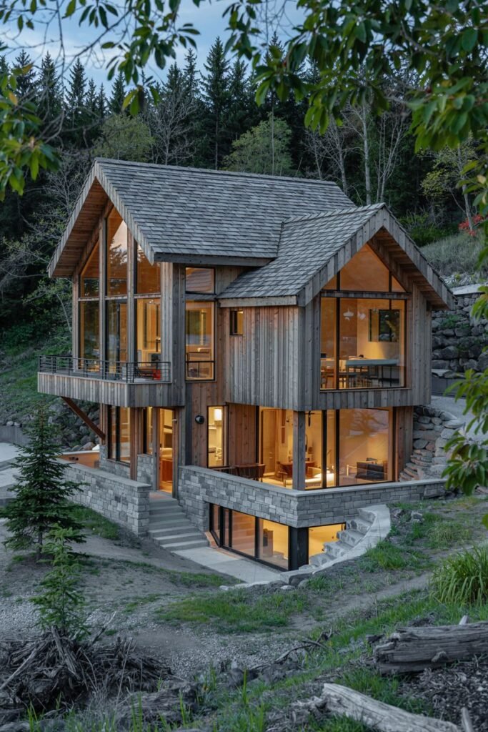 A large, modern rustic house built into a hillside, featuring extensive use of wood siding, stone, and large windows. The design emphasizes connection with the surrounding forest landscape. While large, the natural materials give it a cozy lodge feel against the backdrop of the woods.