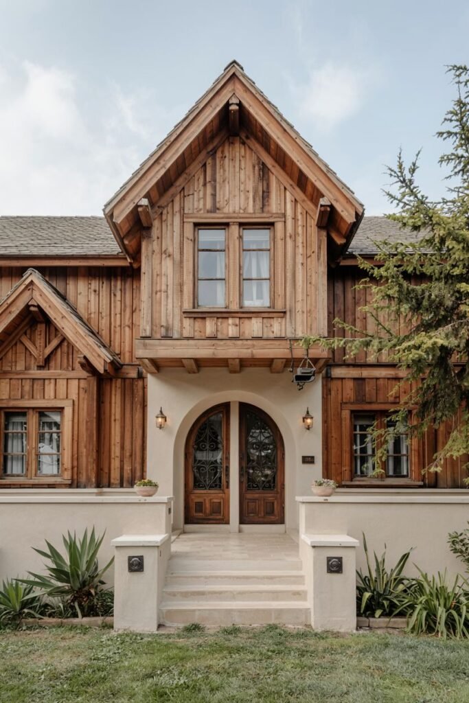 A rustic wooden house with a striking arched double door entryway set within a lighter-colored facade. The upper part of the house features wood siding and windows within a gable. The wooden textures and warm lighting create a cozy welcome.