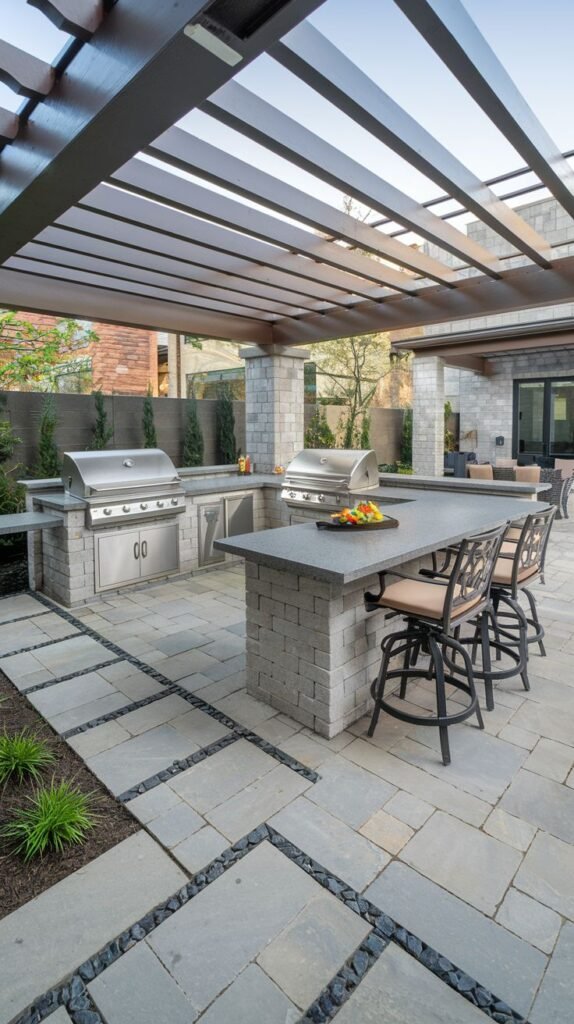 An Outdoor Grill Station featuring two stainless steel grills, a granite countertop, and stone block base, situated under a dark brown pergola. There is a built-in counter with seating at bar stools opposite the grills. The patio is made of large square grey pavers with dark river stones in between them.