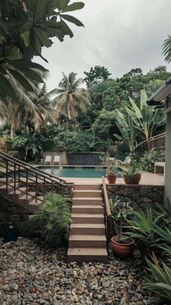 A multi-level pool area is accessed by stone steps leading down from a stone-faced wall. The pool itself is rectangular and surrounded by lush, tropical plants including large-leaved varieties and banana trees. Outdoor chairs are visible by the pool.