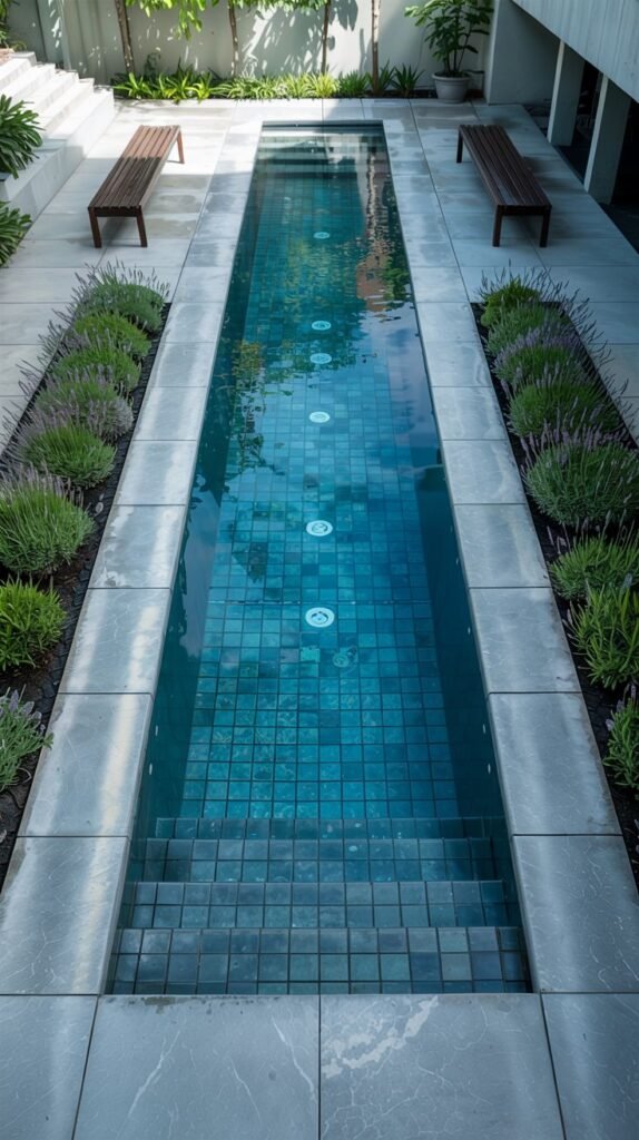 A long, narrow rectangular pool with blue tiled floor is bordered by grey stone coping and beds of lavender plants. Steps lead down into the pool at one end. Two wooden benches are placed on the stone patio area on either side of the pool.