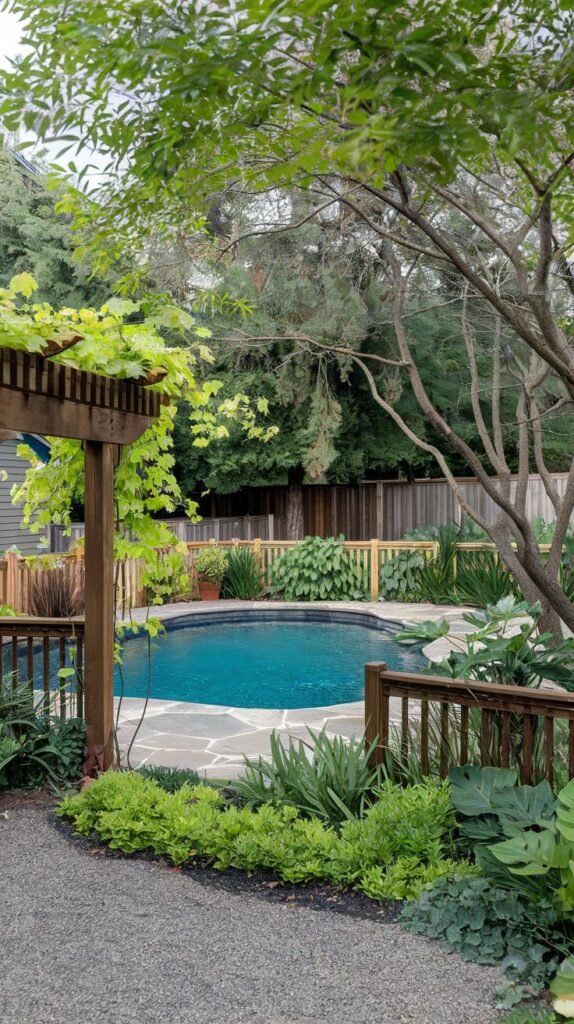 A dark-bottomed pool with stone coping is nestled in a lush backyard. A wooden pergola covered in vines stands in the foreground, and a gravel path leads toward the pool area, which features a wooden fence and various plants.