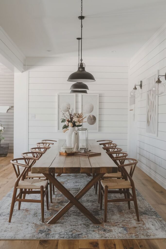 A modern farmhouse dining room with a long wooden table, chairs with woven seats, large pendant lights, and white horizontal plank walls.
