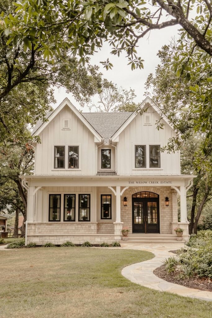 A symmetrical white modern farmhouse with a stone base, dark-framed windows, a gabled roof, and a prominent porch, framed by trees. This appears to be "THE WILLOW CREEK ESTATE".