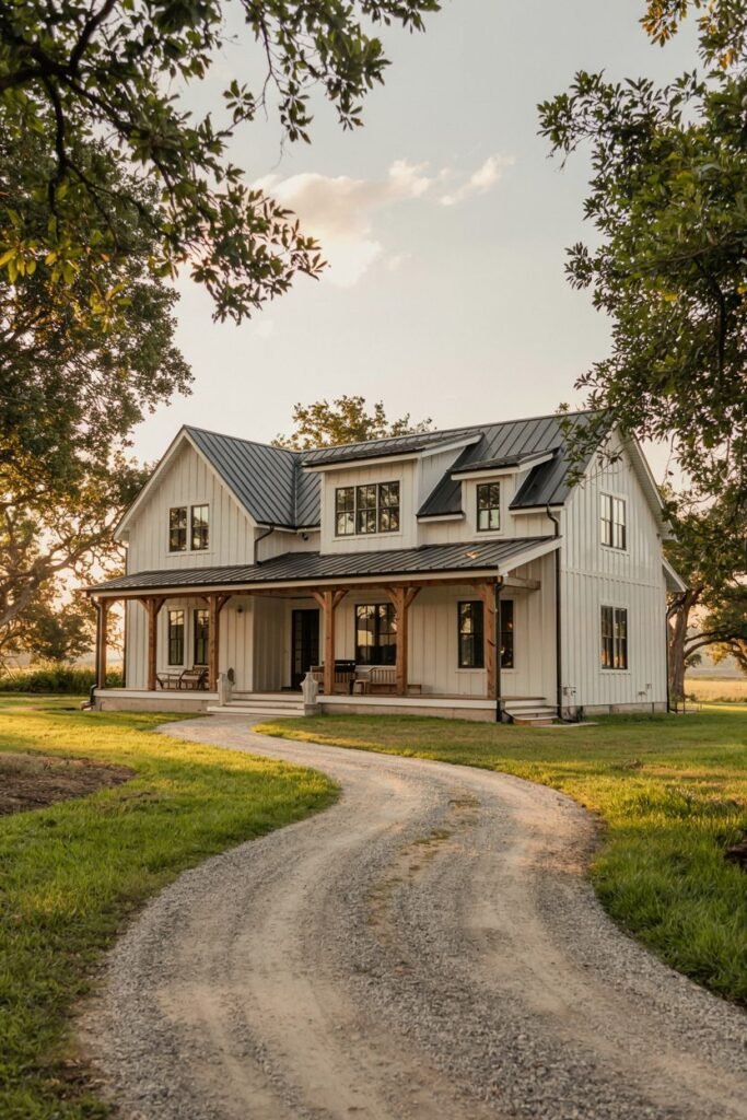 A white-sided two-story modern farmhouse with a dark metal roof, dark windows, and a covered porch supported by natural wood columns, viewed from a gravel driveway under trees.