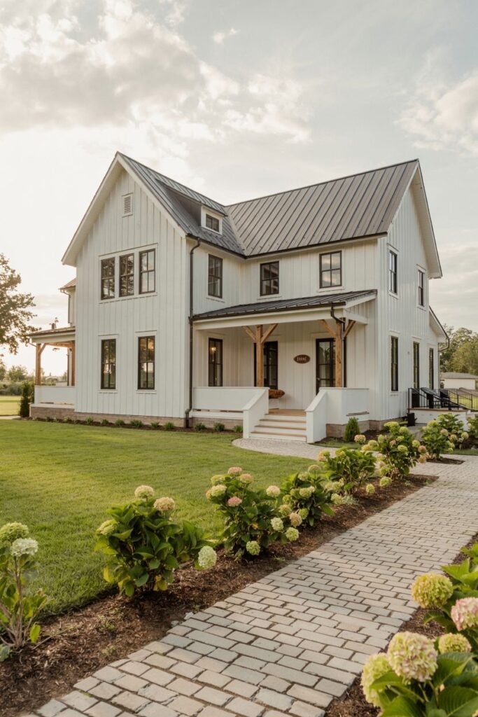 A white-sided two-story modern farmhouse with board-and-batten siding, a dark metal roof, and dark windows, featuring a front path paved with light-colored bricks and bordered by hydrangea plants.
