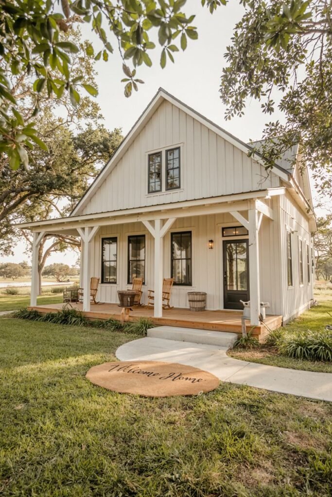 A white-sided modern farmhouse with a low-pitched gabled roof, dark windows, and a welcoming wooden porch with rocking chairs, viewed from a path in the grass with a circular mat saying "Welcome Home".