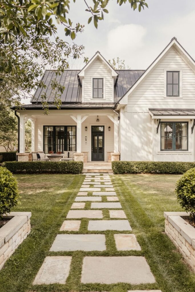 A white-sided modern farmhouse with a dark metal roof, dark windows, and a covered patio/porch with dark furniture, accessed via a stone path laid in the grass.