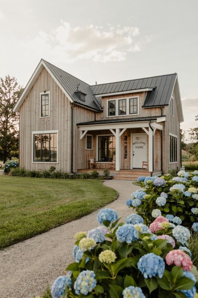 A natural wood-sided two-story modern farmhouse with a dark metal roof, dark windows, and a covered porch with a sign saying "WELCOME HOME", viewed from a gravel path next to large blue and pink hydrangea bushes.