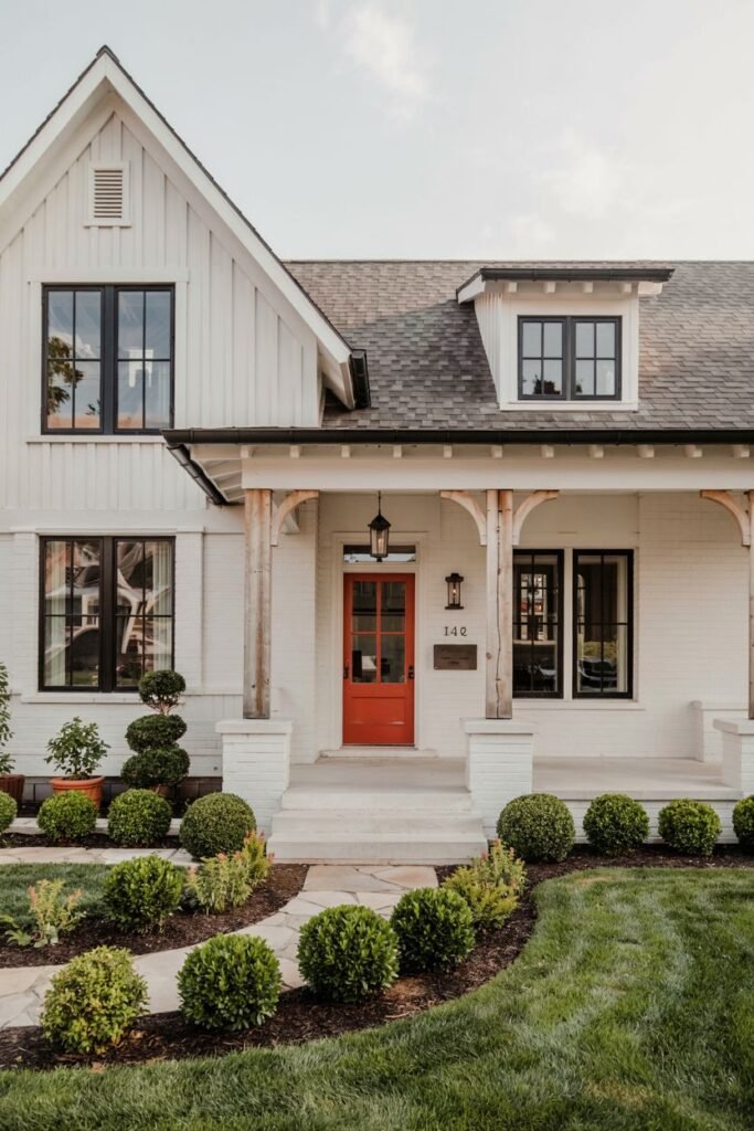 A white brick modern farmhouse with a red front door, dark windows, natural wood accents on the porch structure, and a winding stone path bordered by trimmed bushes.