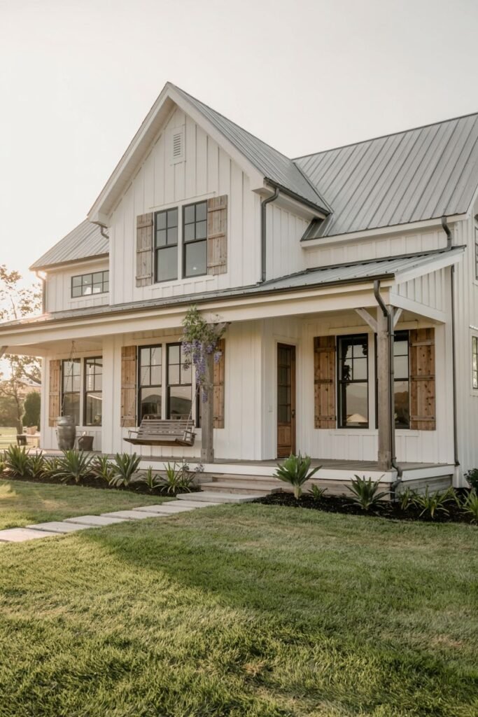 A white-sided modern farmhouse with a light grey metal roof, dark windows with rustic wooden shutters, and a covered porch featuring a porch swing.
