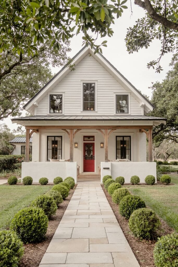A smaller white modern farmhouse featuring a red front door, dark windows, natural wood porch columns, and a walkway lined with trimmed bushes.