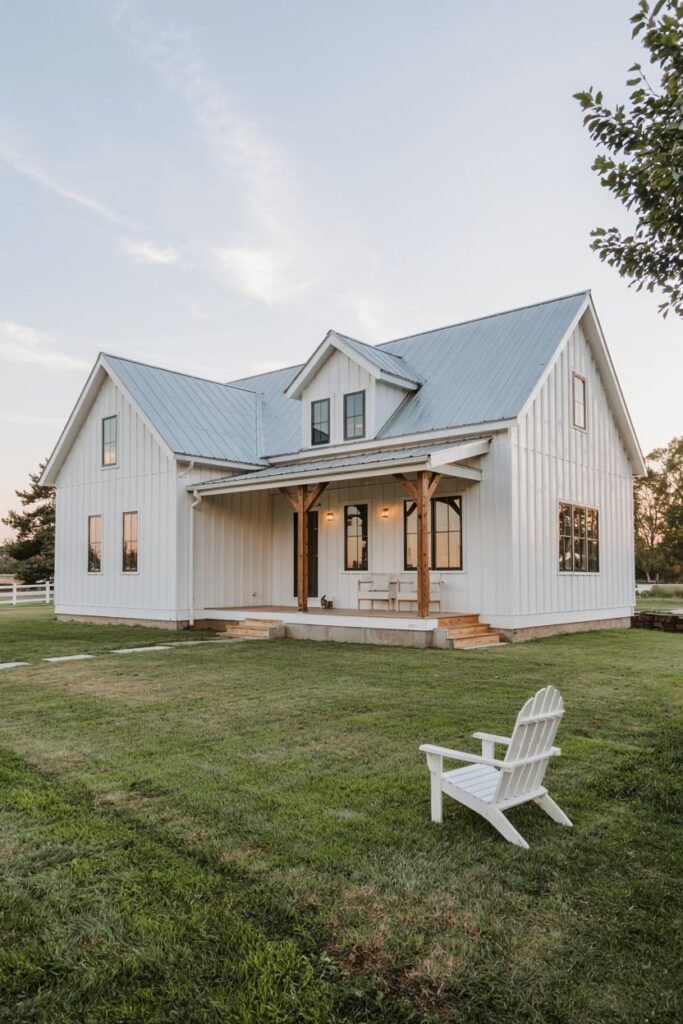 A white-sided two-story modern farmhouse with a light grey metal roof, dark windows, and a covered porch supported by natural wood posts, set in a grassy field with a white Adirondack chair in the foreground.