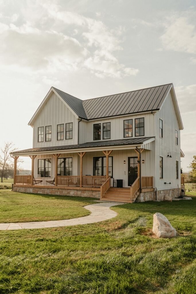 A white two-story modern farmhouse with board-and-batten siding, a dark metal roof, dark windows, and a large wooden deck/porch, situated in a grassy field with a winding path.