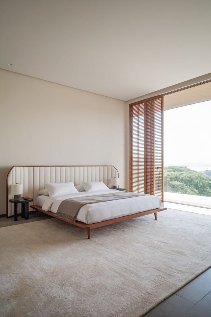 Expansive modern Japandi bedroom showcasing a low-profile bed with a padded headboard, a large rug, and a panoramic view through floor-to-ceiling glass doors with wooden slatted blinds.