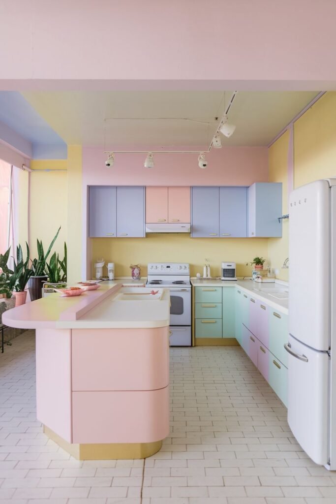 Vibrant pastel kitchen with a mix of colors including pink, yellow, light blue, and light green cabinets and walls. A pink kitchen island with a curved countertop is prominent.