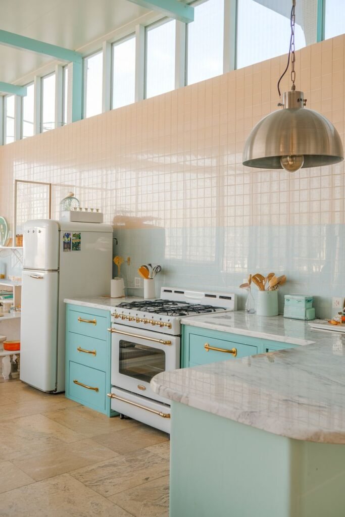 Spacious pastel kitchen with light blue base cabinets, a marble-topped island, and a white refrigerator and range. The backsplash features horizontal bands of pastel pink and light blue tile.
