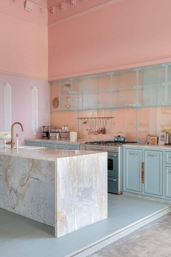 Artistic pastel kitchen with light blue base cabinets, a massive marble kitchen island, and walls painted in shades of pink and light blue. Glass-front cabinets run along the top wall.