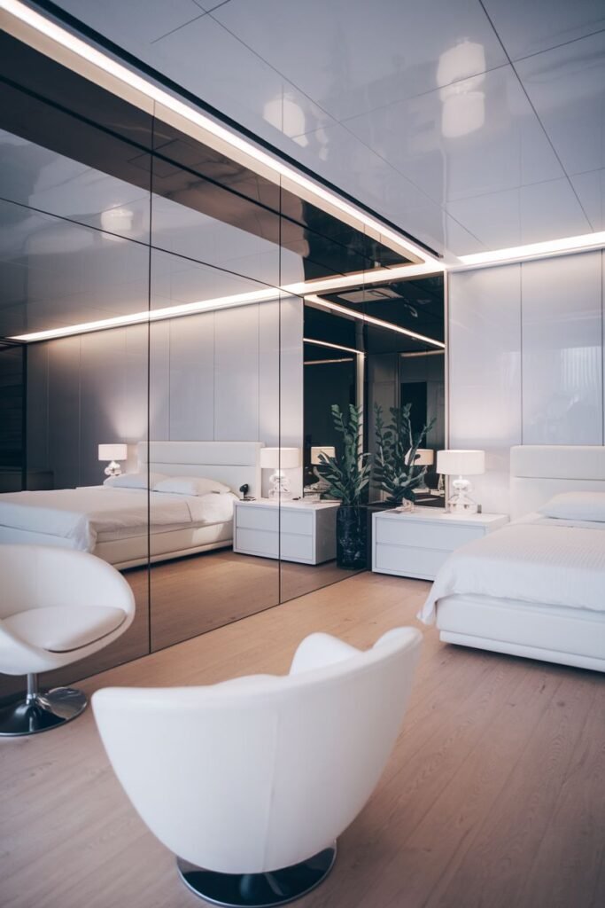 A large mirrored wall composed of rectangular panels with horizontal strip lighting above, reflecting a modern bedroom with two beds and white chairs.