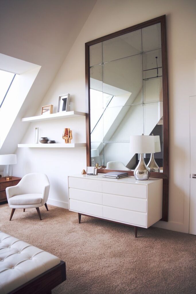A large rectangular mirror with a dark wood frame and grid pattern, placed above a long white dresser in a room with slanted ceilings, shelves, an armchair, and a bench.