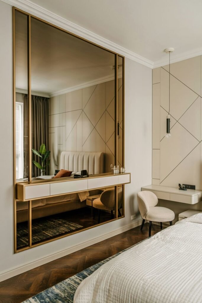 A large rectangular mirror with a gold frame and an integrated shelf with drawers below, mounted on a wall reflecting a bed and room interior, next to a dressing table setup.