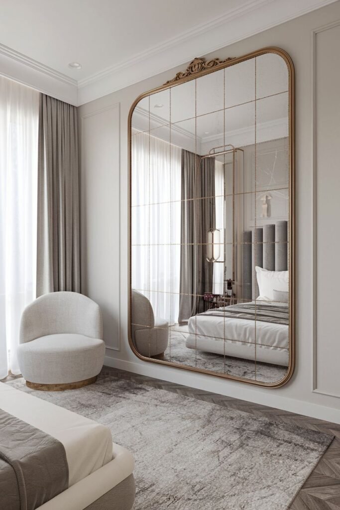 A very large, rectangular gold-framed mirror with a grid pattern, reflecting a bed and bedroom area, mounted on a wall above a white armchair.