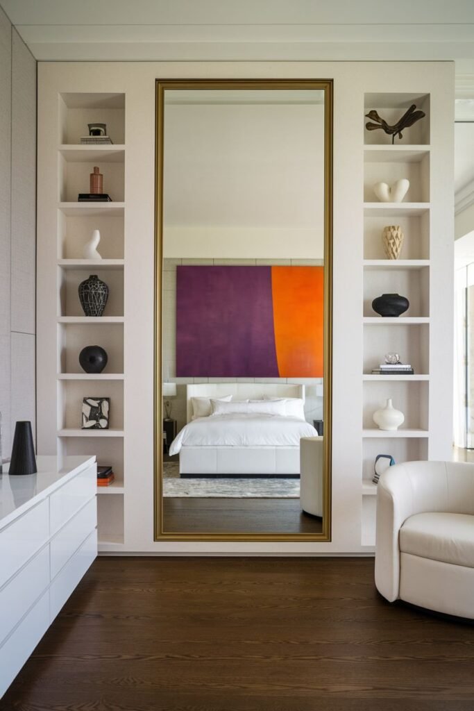 A large rectangular mirror with a gold frame, recessed into a wall between built-in shelves filled with decorative objects, reflecting a bed and a large colorful painting above the bed.