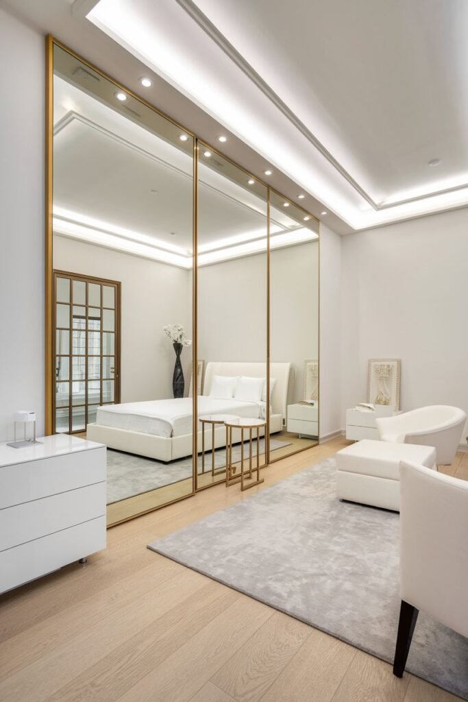 A large wall composed of three tall rectangular mirrored panels with thin gold frames, reflecting a white bedroom with a bed, dresser, and seating area.