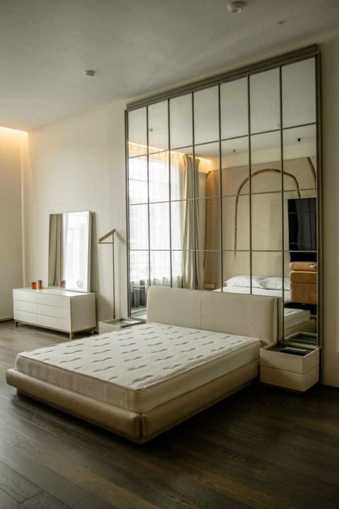 A large mirrored wall composed of square panels, reflecting a bed, dresser, and window in a modern room.