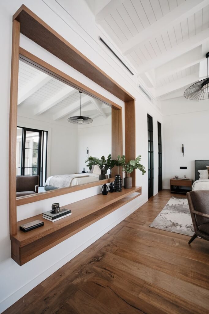 A large rectangular mirror framed by a wide wooden border and integrated shelf, mounted on a white wall in a modern room with a view of another room and wooden floors.