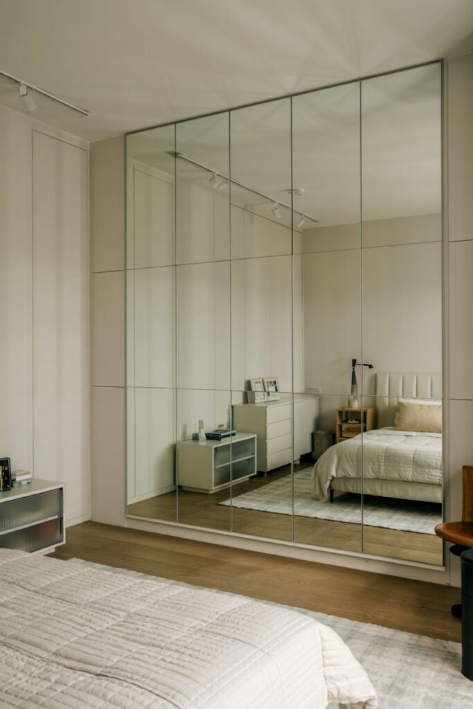 A large mirrored wall composed of rectangular panels, reflecting a bedroom with a bed, dresser, and closet.