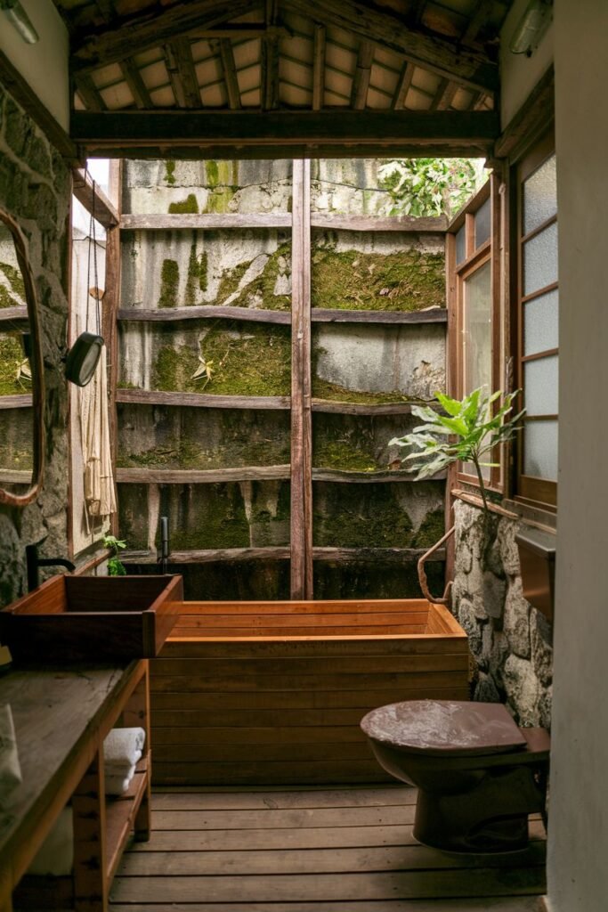 A bathroom with a wooden bathtub, a wooden vanity with a dark sink, a toilet with a brown seat, wooden flooring, rock walls, and a large open window looking out to overgrown greenery.