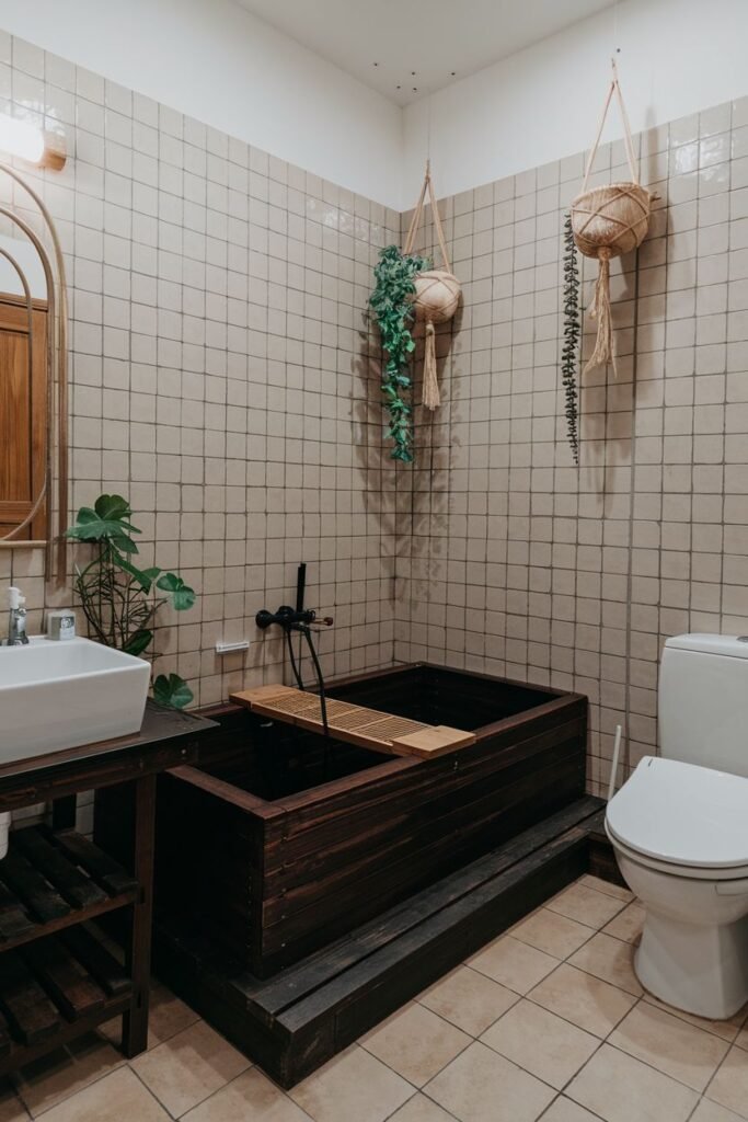 A bathroom with a dark wooden bathtub raised on a platform, a white rectangular sink on a wooden stand, a white toilet, tiled walls, and hanging plants.