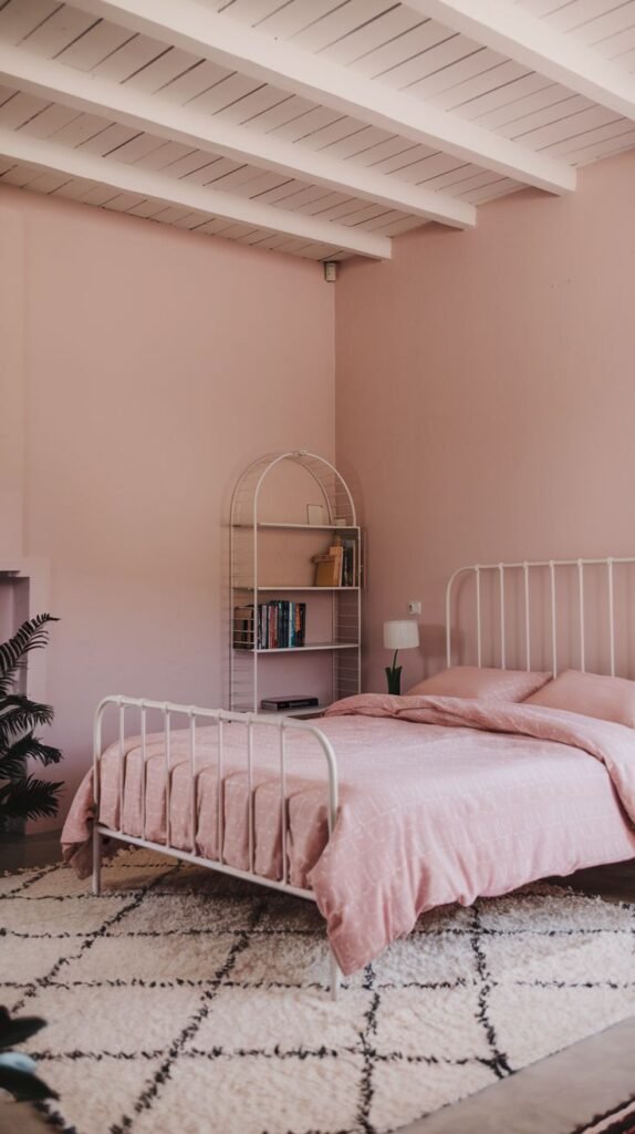 A minimalist yet amazing and comfy pink bedroom, featuring a white metal bed frame with soft pink bedding, a white open shelving unit, and exposed white ceiling beams. A shaggy white rug with subtle black patterns adds texture and warmth to the light-filled space.