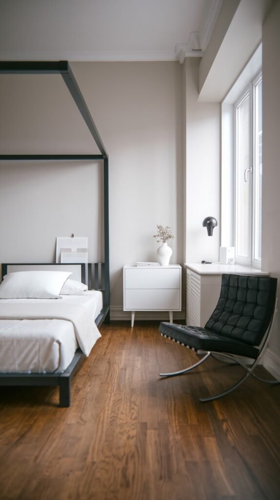 A Contemporary Single bedroom with a black four-poster bed frame, a white nightstand, and a black leather lounge chair. A large window brings in ample light, and the room features wood flooring, offering a chic, minimalist design.