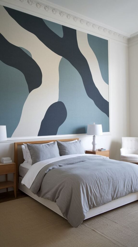 A large abstract mural on a white bedroom wall, featuring organic wavy shapes in muted blues, beige, and white within a framed section. A wooden bed with grey bedding and two white lamps are visible.