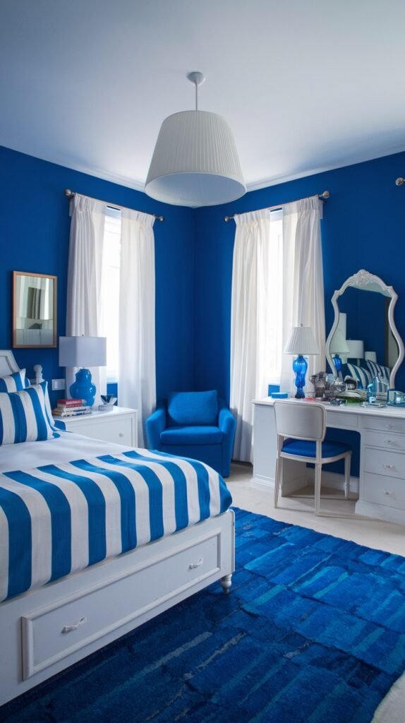 A bright and cozy bedroom with vibrant blue walls, white furniture including a bed and a vanity desk, and a bold blue and white striped comforter. A deep blue rug and a matching blue armchair complete the color scheme, creating a cohesive and cozy space.
